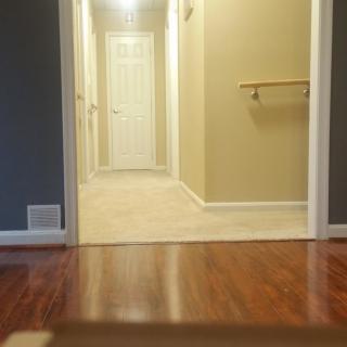 A hallway with a wooden floor leading to a closed door, featuring a handrail and illuminated by ceiling lights.
