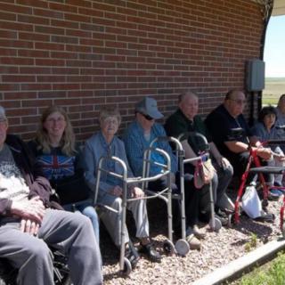 A group of elderly people sitting outside against a brick wall, using walkers.