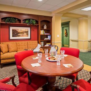 A cozy meeting room with a round table, red chairs, and a comfortable couch, featuring artwork and a bookshelf.