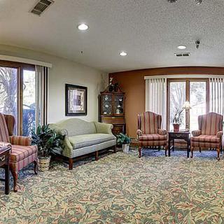 A cozy living room with a sofa, several chairs, and large windows, decorated with patterned carpet and soft lighting.
