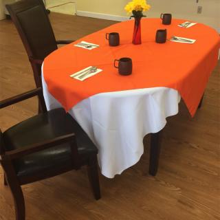 A dining table is set with an orange and white tablecloth, featuring black mugs and a floral centerpiece.