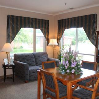 A cozy living room with a sofa, table, and chairs, adorned with flowers and illuminated by lamps near large windows.