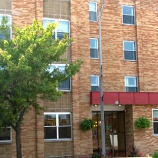 A brick apartment building with multiple windows and a small entrance canopy, surrounded by greenery.