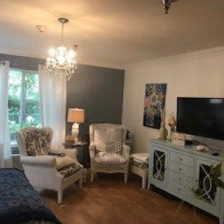 A cozy living room with elegant furniture and a chandelier, featuring armchairs, a dresser with a TV, and warm lighting.