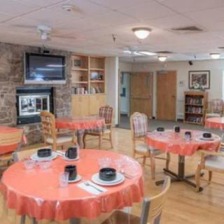 A cozy dining area with tables covered in orange tablecloths, set with bowls and cutlery. The room features a fireplace, a television, and library shelves filled with books.