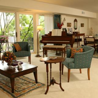 A cozy living room with elegant furniture and a grand piano, featuring natural light from large windows.