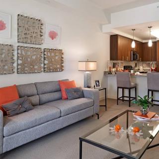 A modern living room with a light gray sofa, orange accents, and an open kitchen area.