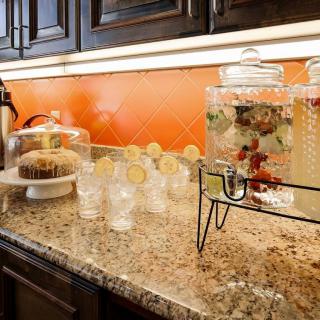 A kitchen counter featuring a beverage dispenser with infused water, glasses garnished with lemon slices, and a cake under a glass dome.