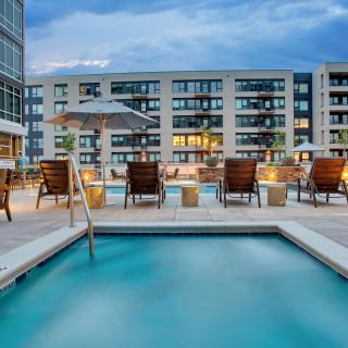 A rooftop swimming pool area with patio chairs and umbrellas in an apartment complex during the evening.