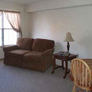 A cozy living room with a brown sofa, side table, and lamp near a window with light curtains.