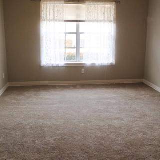 An empty room with beige carpeting and a window covered by sheer curtains.