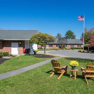 A peaceful motel courtyard with chairs and a table set on a grassy lawn, featuring manicured landscaping and a flag flying high.