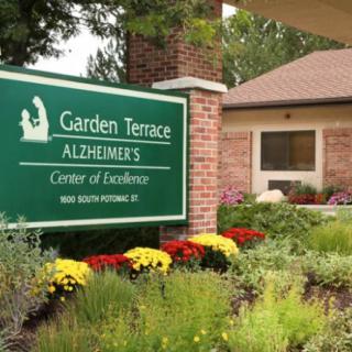 A sign for the Garden Terrace Alzheimer's Center of Excellence in front of a building surrounded by flowers and greenery.