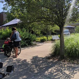 A caregiver pushes a wheelchair-bound person along a scenic path in a park.
