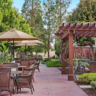A serene outdoor patio with tables and chairs under umbrellas, surrounded by lush greenery and a wooden pergola.