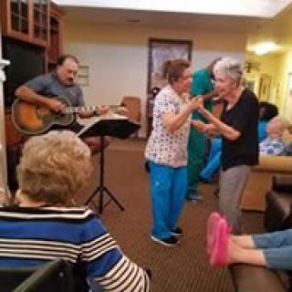 Elderly residents enjoying live music and dancing in a cozy common area.