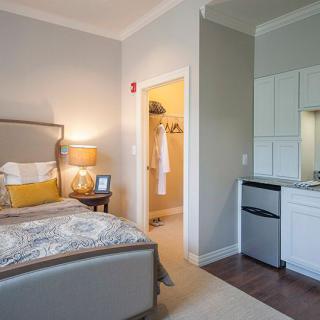 A cozy bedroom with a modern design, featuring a comfortable bed, nightstand with a lamp, and a small kitchenette.