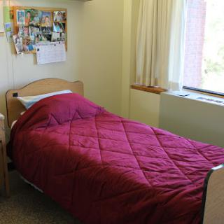 A cozy dorm room featuring a single bed with a red blanket and a bulletin board adorned with photos.