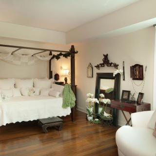 A cozy bedroom with a canopy bed and vintage decor, featuring a wooden floor, an armchair, and a mirror.