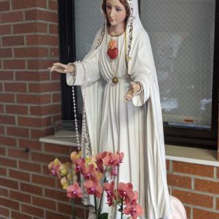 A serene statue of Mary adorned with a rosary, surrounded by vibrant flowers against a brick backdrop.