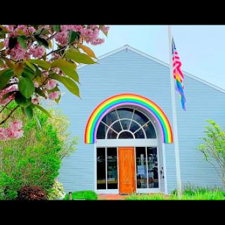 A vibrant community building adorned with a rainbow arch and flags, surrounded by blooming trees.
