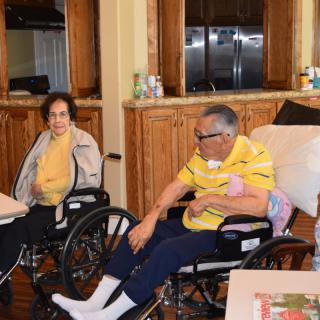 Two elderly individuals sitting in wheelchairs, engaging in conversation in a home setting.