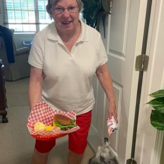 A woman in red shorts and a white shirt is smiling while holding a tray with a burger and chips indoors.