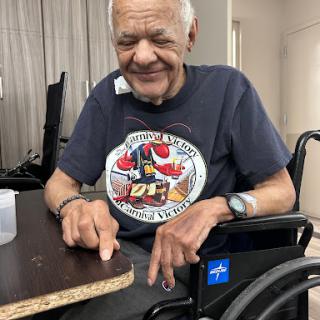 An elderly man in a wheelchair smiles while sitting at a table, wearing a casual t-shirt.