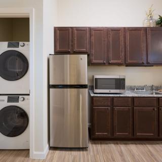 A modern kitchen with a stacked washer and dryer, stainless steel refrigerator, microwave, and brown cabinets.