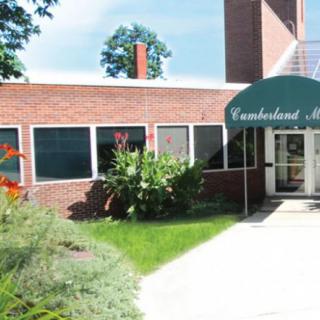 The entrance to a brick church building with an awning, surrounded by greenery and flowers.