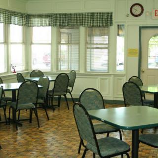 An empty dining area with tables and chairs, illuminated by natural light from the windows.