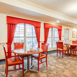 A cozy dining area with red curtains and carpeted flooring, featuring multiple tables and chairs by large windows.