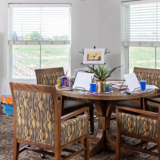 A cozy dining room setup with a round table surrounded by four patterned chairs, featuring notebooks and cups on the table, with natural light streaming in through two large windows.