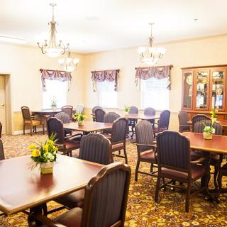 A well-lit dining room with arranged tables and chairs, decorated with chandeliers and floral centerpieces.