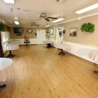 An empty indoor hall with wooden flooring, round tables with white tablecloths, decorative plants, and ceiling fans.