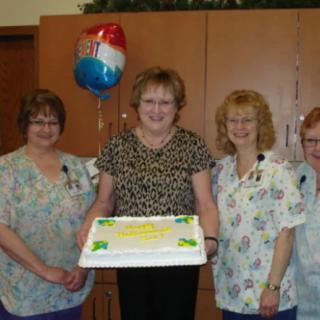 A group of four women celebrate with a cake and balloons indoors.