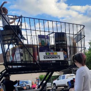 A whimsical moment with a gigantic shopping cart filled with items, as people enjoy the outdoor event.