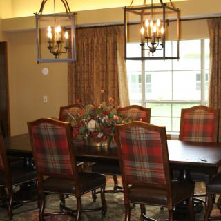 A cozy dining room featuring a large wooden table with plaid upholstered chairs, two elegant light fixtures, and a window adorned with curtains.