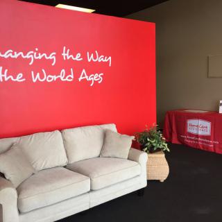 A modern waiting area with a beige couch, red accent wall displaying motivational text, a potted plant, and a table covered with a red tablecloth.