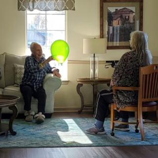 Two seniors are seated in a living room, engaging with a green balloon.