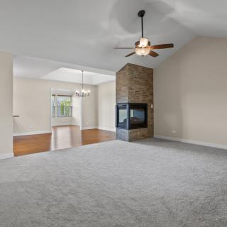 A spacious living room with a central fireplace and ceiling fan, featuring carpeted flooring and ample natural light.