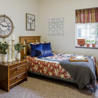 A cozy bedroom with a wooden bed, colorful bedding, and decorative plants. A large wall clock and artwork adorn the walls.