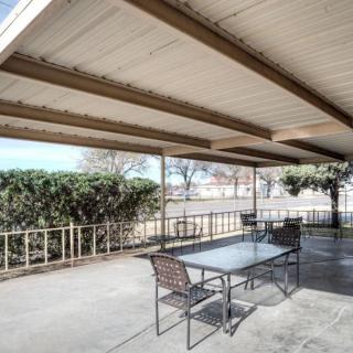 A covered outdoor patio area with a table and chairs, surrounded by landscaping and bushes.