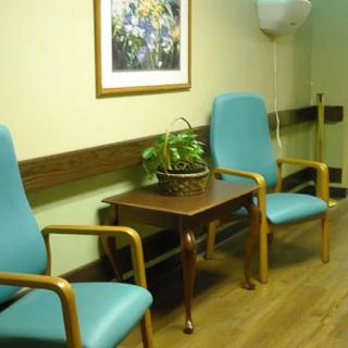 A quiet waiting area with two green chairs, a small table, and a plant under a framed painting.