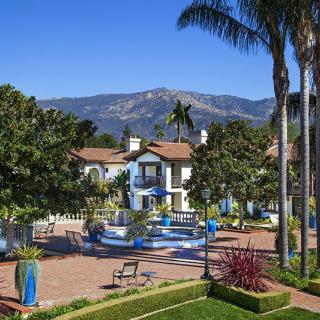 A picturesque resort courtyard with a fountain, surrounded by palm trees and mountains in the background.