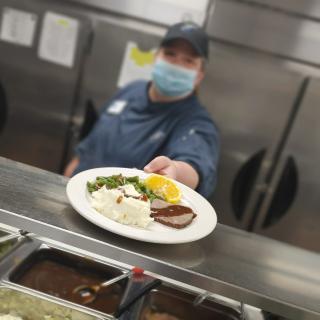 A chef wearing a mask presents a plated meal in a restaurant kitchen.