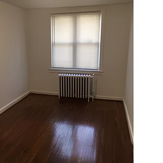 An empty room with hardwood floors, featuring a window with blinds and a radiator underneath.