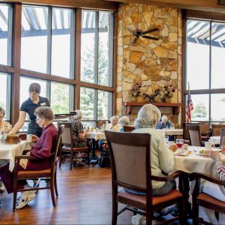 Seniors dining in a well-lit restaurant with large windows and a stone fireplace.