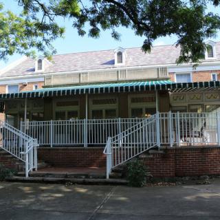 A residential house with a large porch, featuring brick steps and white railings, shaded by trees.