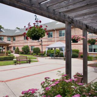A serene courtyard featuring blooming flowers, hanging plants, and benches, surrounded by a welcoming building.